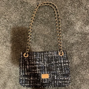 Rutig axelväska från Tiamo med kedja - Snygg axelväska från Tiamo i rutigt tyg med svart, blått och vitt. Väskan har en guldfärgad kedjerem med inslag av svart skinn och ett elegant spänne framtill. Bandet har gått av där uppe men de är inget som märks!!💓💓