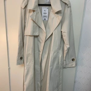 Cremevit  trenchcoat från Bershka S - Snygg  trenchcoat från Bershka i storlek S. Jackan har klassisk krage, långa ärmar, stora fickor och bälte i midjan för en cool siluett. Perfekt för dig som gillar stilrena och tidlösa plagg med modern touch.