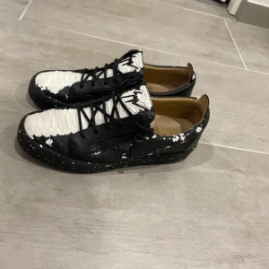 Svarta och vita sneakers från Giuseppe Zanotti - Unika sneakers från Giuseppe Zanotti med svart och vit färg, coolt reptilmönster och stänkmålade detaljer. Skorna har snörning, dragkedja på sidan och rund tå. Tillverkade i skinn med texturerad yta och logga på plösen. Perfekt för dig som gillar statement pieces.