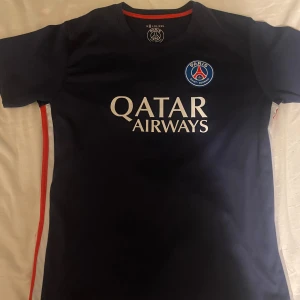 PSG matchtröja blå Qatar Airways - Snygg PSG fotbollströja i mörkblått med klubbmärke på bröstet och Qatar Airways-tryck på framsidan. Röd och vit detalj längs sidorna, rund hals och korta ärmar. Perfekt för dig som vill visa vilket lag du håller på.