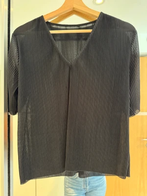 Svart plisserad blus med v-ringning - Säljer en svart plisserad blus med v-ringad hals och korta ärmar. Blusen har en luftig passform och är tillverkad i ett lätt, glansigt syntetmaterial som ger en snygg struktur. Svart ribbad skir v-ringad kortärmad topp. Perfekt till tex julfest och nyår.