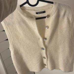 Beige stickad väst från Gina Tricot - Säljer en beige stickad väst från Gina Tricot med rund hals och silvriga knappar framtill. Västen har en mjuk och mysig känsla, perfekt att styla över en t-shirt eller skjorta. Modellen är rak och har ribbade kanter vid hals och nederkant.