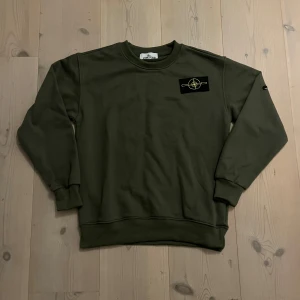 Olivgrön Stone Island sweatshirt - Snygg olivgrön sweatshirt från Stone Island med klassisk patch på ärmen. Tröjan har rund hals, ribbade muddar och är långärmad. Perfekt för dig som gillar streetwear och vill ha en clean look med ikonisk logga.