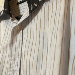 Randig skjorta Selected med svart krage- NY - Vit skjorta från Selected i storlek S med tunna bruna ränder och svart krage med broderade detaljer. Klassisk knappstängning framtill och vid ärmslut. Skjortan har en clean och stilren look, perfekt för dig som gillar detaljer och snygga kontraster.