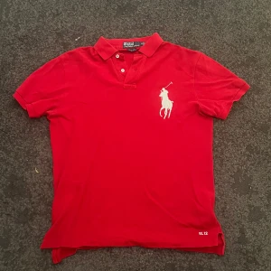Röd pikétröja från Ralph Lauren - Snygg röd pikétröja från Ralph Lauren med klassisk vit polo-spelare broderad på bröstet. Tröjan har krage, korta ärmar och knappar vid halsen. RL 12 broderat nedtill. Perfekt för dig som gillar stilren och sportig look.