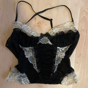 Svart korsettopp med spetsdetaljer - Säljer en svart korsett med tunna axelband och snygga ljusa spetsdetaljer. Axelbanden korsas i ryggen. Toppen har en liten rosett framtill och genomskinliga partier i spetsen. 