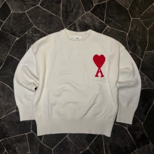 Ami Paris Stickad - Vit stickad tröja från Ami med rund hals och ribbade muddar. På bröstet finns ett stort rött hjärta med bokstaven A under, vilket ger en unik och snygg detalj. Perfekt för dig som gillar stilrena plagg med en twist.