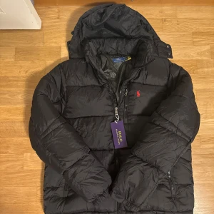 Svart pufferjacka från Polo Ralph Lauren - Svart pufferjacka från Polo Ralph Lauren med röd broderad logga på bröstet. Jackan har huva, två sidofickor med dragkedja och en kraftig dragkedja framtill. Materialet är syntet och jackan har en quiltad design som ger extra volym och värme.