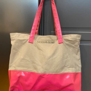 Victoria's Secret shoppingväska beige/rosa - Stor shoppingväska från Victoria's Secret i beige canvas med glansig rosa botten och matchande rosa handtag. Väskan har ett rymligt fack och logga i silver framtill. Perfekt för att bära med sig allt man behöver till skolan eller stan. 38 cm bred 33 cm hög 17 cm djup