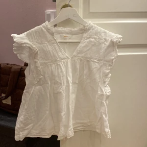 Vit blus med volang från Gina Tricot - Supersöt vit blus från Gina Tricot med volangdetaljer vid ärmar och hals. Blusen har v-ringning och är tillverkad i ett lätt och luftigt bomullstyg, perfekt för varmare dagar. Den är ärmlös och har en avslappnad passform som ger en skön känsla.