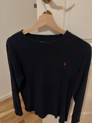 Mörkblå långärmad tröja från Polo Ralph Lauren - Säljer en stilren mörkblå långärmad tröja från Polo Ralph Lauren med det klassiska röda logotypbroderiet på bröstet. Perfekt för en avslappnad och snygg look. Tröjan är tillverkad i ett mjukt och bekvämt material.