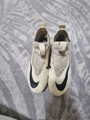 Nike Mercurial Air Zoom fotbollsskor - Nike Mercurial Air Zoom fotbollsskor i beige med svarta detaljer och stor Swoosh-logga på ovansidan. Skorna har en strömlinjeformad design, elastisk ovandel och dragflik bak för enkel påtagning. Tillverkade i syntetmaterial och textil för bra passform på planen.