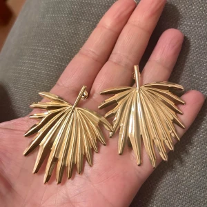 Guldiga örhängen i palmblad-design - Säljer ett par örhängen i guldigt metallmaterial formade som palmblad. De har en cool och unik design med blanka ytor och tydliga detaljer som ger en tropisk vibe. Perfekta för att lyfta din stil och sticka ut i mängden.