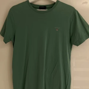 Grön t-shirt från GANT, storlek S - Snygg grön t-shirt från GANT i storlek S med klassisk rund hals och diskret logga broderad på bröstet. Modellen är regular fit och tillverkad i mjuk bomull som känns skön mot huden. Perfekt för dig som gillar stilrena och enkla plagg.