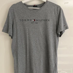 Grå t-shirt Tommy Hilfiger - Snygg grå t-shirt från Tommy Hilfiger med tryck på bröstet och liten logga på ärmen. Tillverkad i mjuk ekologisk bomull, rund hals och klassisk passform. Perfekt för dig som gillar stilrena och enkla plagg med ikonisk branding.