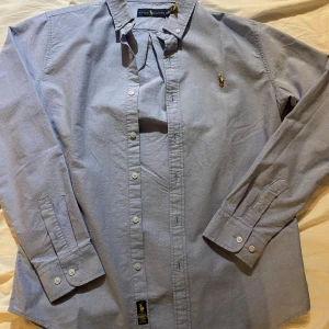 Blå skjorta från Ralph Lauren - Snygg ljusblå skjorta från Ralph Lauren med klassisk button-down krage och broderad logga på bröstet. Skjortan har långa ärmar, knappar framtill och är gjord i mjuk bomull. Perfekt för dig som gillar stilrena och tidlösa plagg.