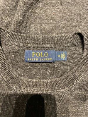 Mörkgrå tröja från Polo Ralph Lauren - Mörkgrå tröja från Polo Ralph Lauren med klassisk rund hals och diskret broderad logga på bröstet. Tröjan är långärmad och har ribbade muddar vid ärmslut och nederkant. Perfekt för dig som gillar stilren och enkel design.