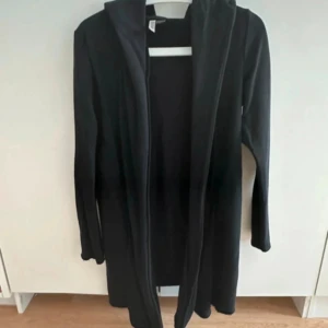 Svart lång cardigan jacka med huva - Svart lång cardigan jacka från DIVIDED med huva och öppet framtill. Jackan är i mjukt tyg, har långa ärmar och en loose passform. Perfekt för lager-på-lager och chill stil. Enkel design utan mönster eller detaljer