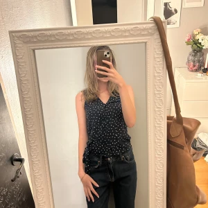 Mörkblå prickig linnetopp med knyt - Säljer en mörkblå ärmlös linnetopp med vita prickar och v-ringning. Toppen har ett snyggt knyt i sidan som ger en cool detalj. Perfekt till jeans eller kjol för en fräsch look. 