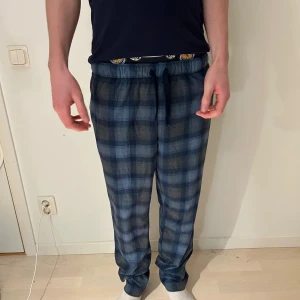 Blårutiga mjukisbyxor med snörning - Säljer ett par blårutiga mjukisbyxor med snörning i midjan. Byxorna har en relaxed passform och elastisk midja för extra komfort. Perfekta för chill dagar hemma eller när du vill vara extra bekväm. Mönstret är klassiskt rutig i olika blå toner.