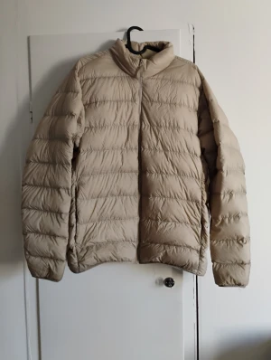 Beige dunjacka från Uniqlo - Säljer en beige dunjacka från Uniqlo Ultra Light Down. Jackan har en clean look med ståkrage, horisontella sömmar och dragkedja framtill. Materialet är nylon och polyester, vilket gör den lätt och smidig att bära. Perfekt för kyliga dagar.