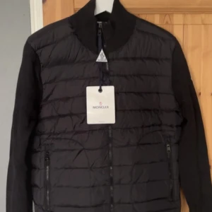 Svart pufferjacka från Moncler - Svart pufferjacka från Moncler med quiltad framsida och stickade ärmar. Jackan har hög krage, två sidofickor med dragkedja och Moncler-logga på ärmen. Materialmix av polyester och stickad textil, perfekt för kyliga dagar. Snygg och stilren design med hel dragkedja framtill.