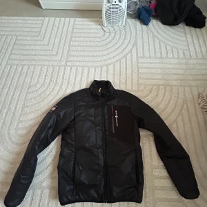 Svart pufferjacka från Sail Racing - Svart pufferjacka från Sail Racing med USA 77 Extreme Sailing patch på ärmen och Sail Racing Link Technology. Jackan har lång ärm, dragkedja framtill och en bröstficka med logga. Perfekt för dig som gillar sportig och clean stil. Och den är limited edition 2017 model 