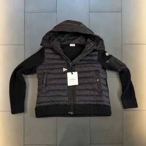 Svart Moncler pufferjacka med huva - Svart pufferjacka från Moncler med stickade ärmar och huva. Jackan har ribbade muddar, två sidofickor med dragkedja och en extra ficka med dragkedja på ärmen. Materialmix av polyester och stickad ull ger en cool look. Perfekt för höst och vinter.