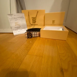 Burberry bälte  - Snyggt Burberry bälte i skinn med det ikoniska beige, svarta och röda rutmönstret. Bältet har ett stort guldfärgat 'B'-spänne framtill och levereras med originalbox, dustbag och kvitto. Perfekt accessoar för att lyfta din outfit.