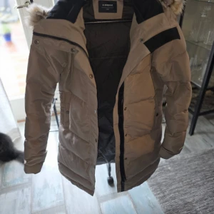 Beige pufferjacka med fuskpäls från Everest. Oanvänd - Snygg beige pufferjacka från Everest med svart detalj på axlarna och huva med fluffig fuskpäls. Jackan har dragkedja och knappar framtill, långa ärmar och quiltad design. Perfekt för kalla dagar och riktigt mysig att bära. Oanvänd 
