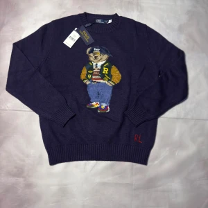 Mörkblå stickad tröja Polo Bear - Mörkblå stickad tröja från Polo Ralph Lauren med ikonisk Polo Bear broderad framtill. Tröjan har rund hals, ribbade muddar och RL-broderi vid nederkant. Materialet är en lyxig blandning av ull och kashmir för extra mjuk känsla.