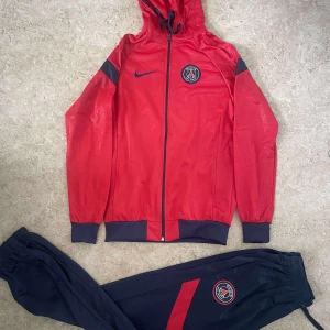 Röd PSG vindjacka från Nike - Röd PSG vindjacka från Nike med huva och hel dragkedja. Jackan har svarta detaljer på ärmar och muddar, samt PSG-logga och Nike-logga på bröstet. Perfekt för dig som älskar fotboll och vill ha en sportig look.