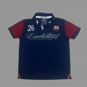 Marinblå pikétröja Vinson Polo Club - Snygg marinblå pikétröja från Vinson Polo Club med röda korta ärmar och broderade detaljer på bröstet, inklusive Union Jack och texten 'Excellence'. Klassisk krage och knappslå framtill. Perfekt för dig som gillar sportig och stilren look.