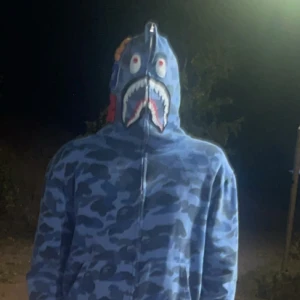 Blå camo hoodie med hajmotiv - Säljer en blå camo hoodie från BAPE med hajmotiv på huvan som täcker hela ansiktet. Hoodien har dragkedja hela vägen upp och coola detaljer i rött och vitt på huvan. Perfekt för dig som vill sticka ut med streetwear.