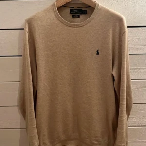 Stickad tröja Polo Ralph Lauren - Snygg beige långärmad tröja från Polo Ralph Lauren i slim fit-modell. Tillverkad av mjuk pima bomull och har den klassiska broderade loggan på bröstet.  Köpt för 1600 kr, säljer för endast 500.Perfekt för dig som gillar stilrena och enkla plagg med exklusiv känsla.