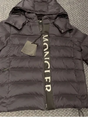 Svart pufferjacka från Moncler - Säljer en svart pufferjacka från Moncler med stor logga längs dragkedjan. Jackan har huva, quiltad design och dragkedja framtill. Perfekt för kalla dagar och har en modern, loose passform som ger en cool vibe.