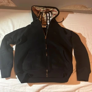 Svart Burberry hoodie med dragkedja - Svart hoodie från Burberry med klassiskt rutigt foder i huvan och broderad logga på bröstet. Tröjan har dragkedja framtill, känguruficka och långa ärmar. Perfekt för dig som vill ha en stilren och lyxig streetwear-look.