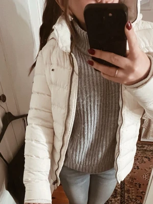 Vit pufferjacka från Tommy Hilfiger - Säljer en snygg vit pufferjacka från Tommy Hilfiger med huva och dragkedja. Jackan har quiltad design, lång ärm och klassisk logga på bröstet och ärmen. Luvan går att ta bort.