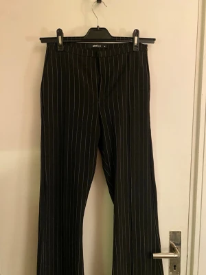 Svarta randiga kostymbyxor Gina Tricot - Svarta kostymbyxor från Gina Tricot i storlek M med smala vita ränder. Byxorna har rak passform, dragkedja och knapp framtill samt bälteshällor. Snyggt klassiskt snitt som funkar till många olika looks.