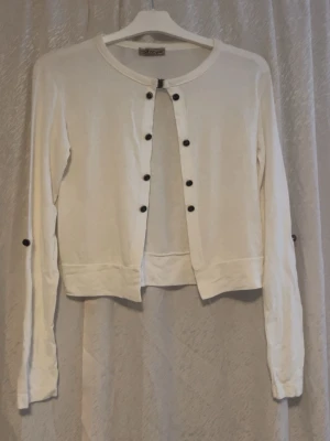 Vit kofta med svarta knappar från Vila - Snygg vit kofta från Vila med rund halsringning och svarta knappar längs hela framsidan. Långärmad modell i mjukt material, perfekt att slänga över en topp. Enkel och stilren design som funkar till det mesta.