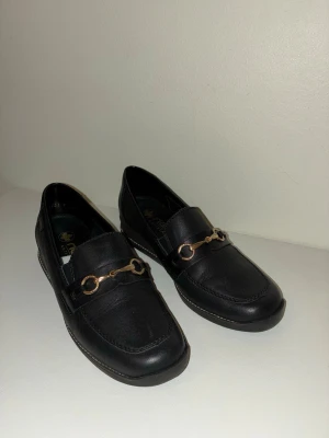 Svarta loafers från Rieker med gulddetalj - Helt nya, oanvända snygga svarta loafers från Rieker med guldfärgad metallspänne över foten. Skorna har rund tå, platt sula och är tillverkade i skinn med diskret logga på sidan. Perfekt för dig som gillar klassisk stil med en modern twist.