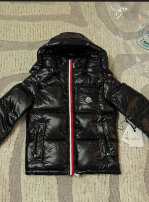 Moncler jacka  - Svart glansig pufferjacka från Moncler med huva, röd och vit rand längs dragkedjan, logga på bröstet och praktiska fickor med dragkedja. Jackan är tillverkad i syntetmaterial och har en quiltad design som ger en cool och sportig vibe. Helt ny aldrig använt. den är strl S tar byten bror på vad det är och fraktar genom plick har tyvär ingen kvitto