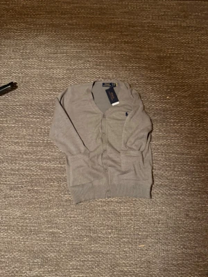 Grå cardigan från Polo Ralph Lauren - Snygg grå kofta från Polo Ralph Lauren med klassisk v-ringning och knappar framtill. Tröjan har långa ärmar, två fickor och en liten broderad logga på bröstet. Mjuk och tunn bomullskvalitet som passar perfekt till lager, på lager. Säljer på grund av att den va för stor