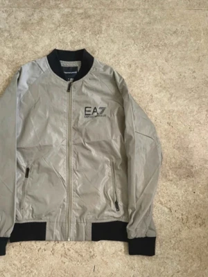 Beige bomberjacka från EA7 Emporio Armani - Snygg beige bomberjacka från EA7 Emporio Armani med svarta muddar och krage. Jackan har dragkedja framtill, två fickor med dragkedja och logga på bröstet. Perfekt för dig som vill ha en stilren och modern look.