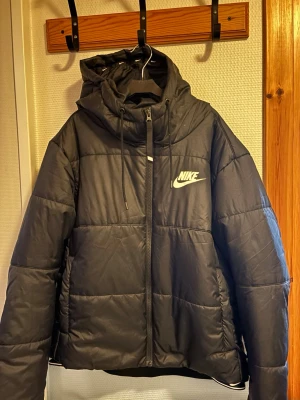 Svart pufferjacka från Nike - Svart pufferjacka från Nike med huva och snörning. Jackan har en quiltad design, dragkedja framtill och Nike-logga på bröstet. Insidan av huvan har små vita Nike-loggor. Perfekt för kalla dagar och har en loose passform.