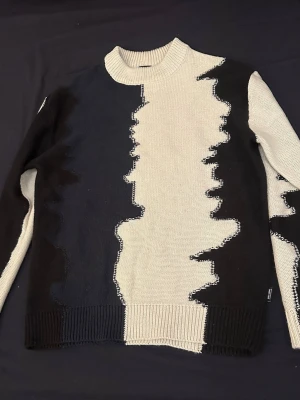 Svart och vit stickad tröja med mönster från only & sons - Säljer en cool stickad tröja i svart och vit med ett unikt, vågigt mönster som går över hela plagget. Tröjan har ribbad hals, muddar och nederkant samt långärmad passform. Perfekt för dig som gillar grafiska detaljer och vill sticka ut lite extra.