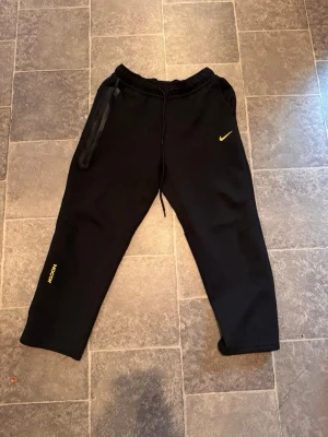 Nike NOCTA svarta mjukisbyxor M - Svarta mjukisbyxor från Nike NOCTA med gul Swoosh och NOCTA-tryck. Byxorna har snörning i midjan, tapered passform och detaljerade sömmar. Materialet är mjukt och skönt, perfekt för chill eller träning. Flera små tryck i gult på benen ger en unik vibe.