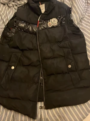 Svart dunväst från Moncler - Säljer en svart dunväst från Moncler med ikonisk logga på bröstet och tryckknappar på fickorna. Västen har quiltad design och dragkedja framtill. Insidan är fodrad med Moncler-tryck och etiketter. Perfekt för dig som vill ha en snygg och varm look.