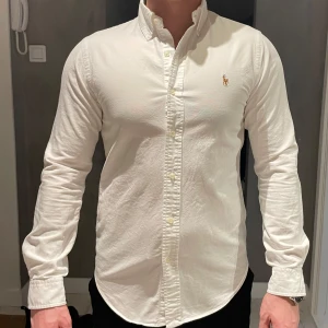 Vit skjorta från Ralph Lauren - Klassisk vit Oxford skjorta från Ralph Lauren med button-down krage och broderad logga på bröstet. Skjortan har smal passform och är tillverkad i mjuk bomull. Utmärkt skick! 
