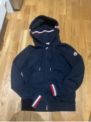 Moncler zip hoodie -   "Navyblå Moncler hooded sweatshirt med hel dragkedja. Stilrena vita och röda ränder på huva och muddar ger en sportig touch. Moncler-logga på vänster ärm. Perfekt för både vardag och sportiga tillfällen. Mycket bra skick!" 